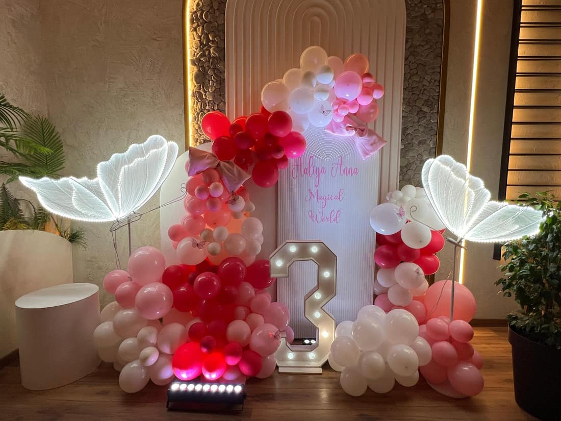 Third birthday backdrop with red, pink and white balloons, a light-up 3 and neon butterfly wings