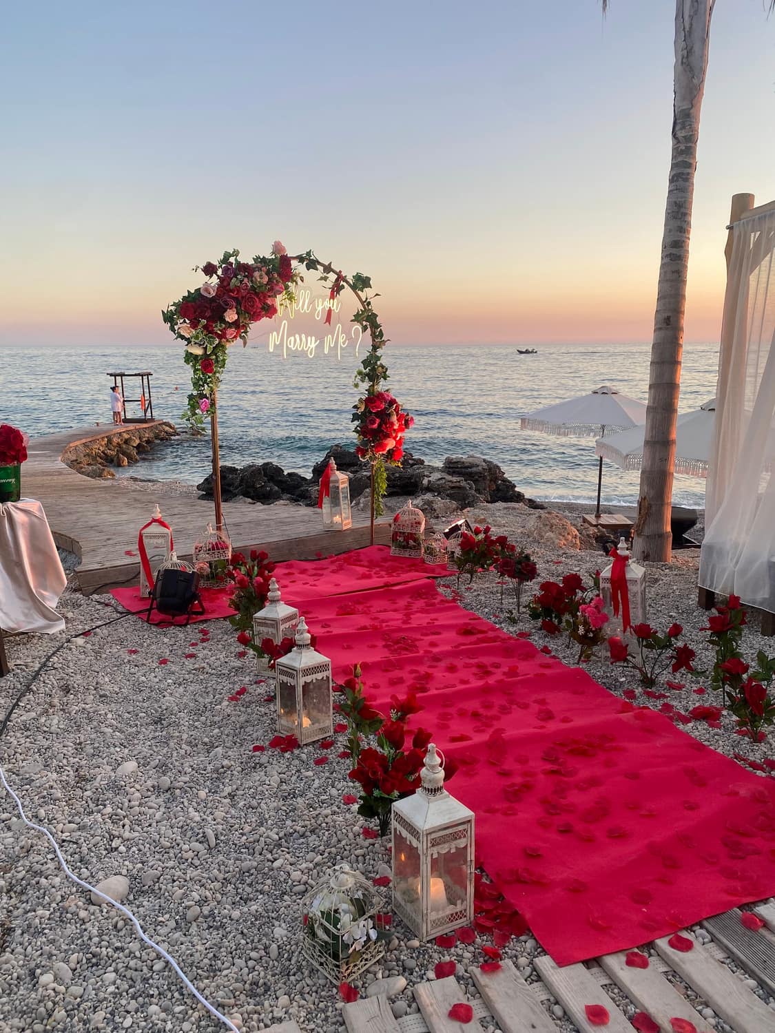 Seaside wedding ceremony at sunset: a red aisle lined with lanterns leading to a rose arch by the water