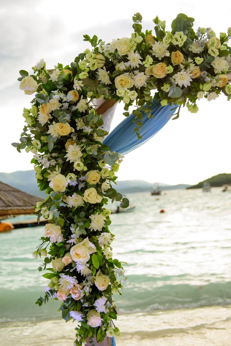 White and yellow flower arch on a beach with boats on the water