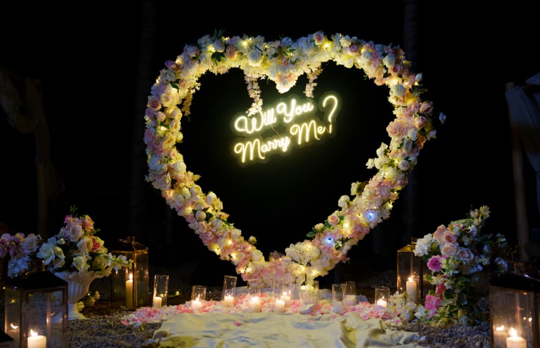 Flower heart with a neon proposal message, lanterns and candles around it at night