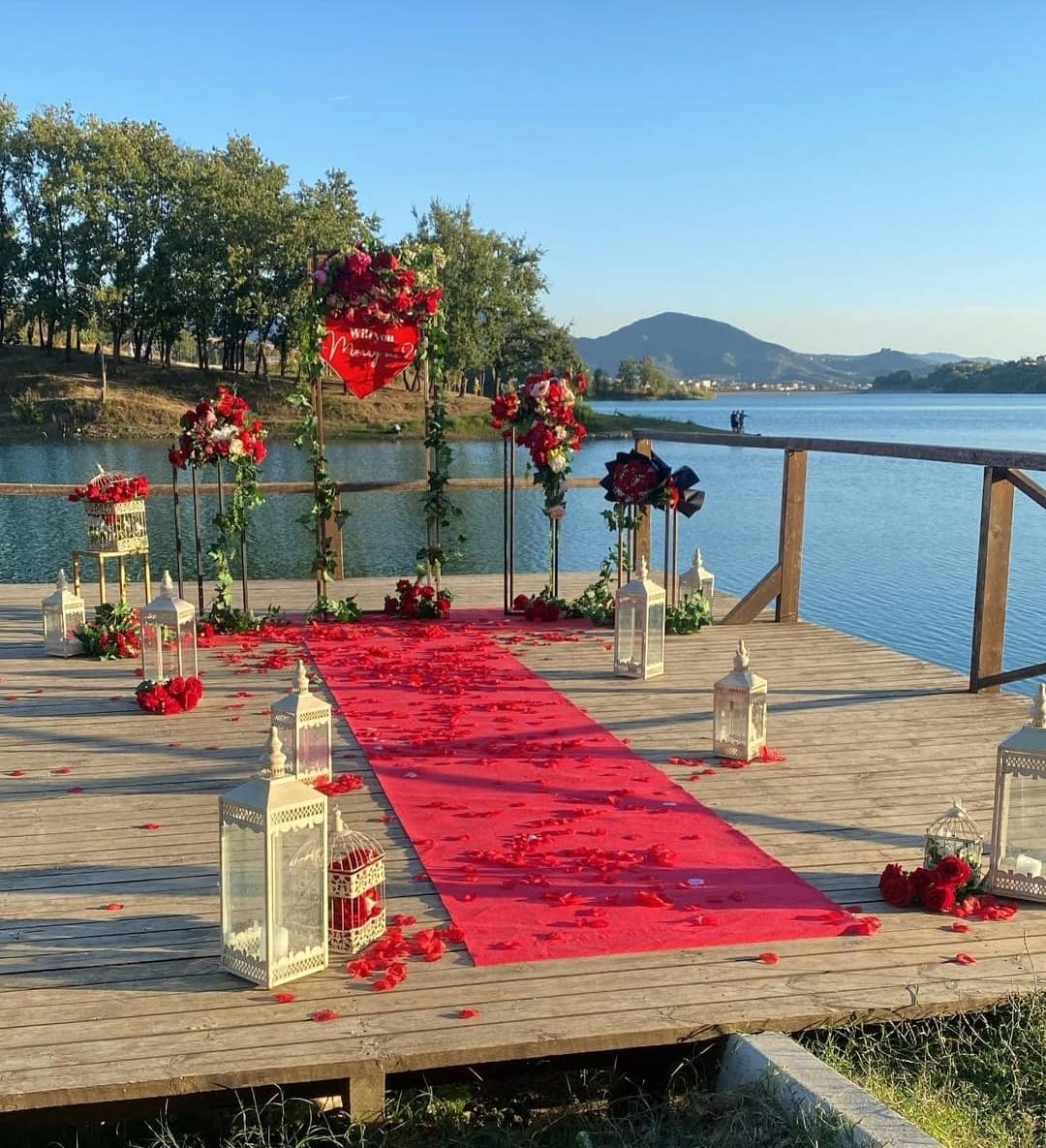 Ceremony on a wooden pier by a lake: red aisle, rose arch and white lanterns