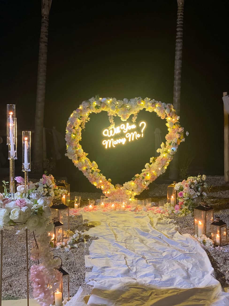 Proposal setup at night: a flower heart lit with 'Will you marry me?' in neon, candles on the ground