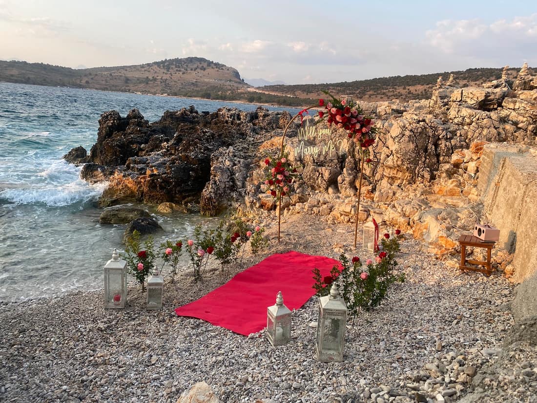 Proposal setup in a rocky cove: a red carpet, roses and lanterns by the sea