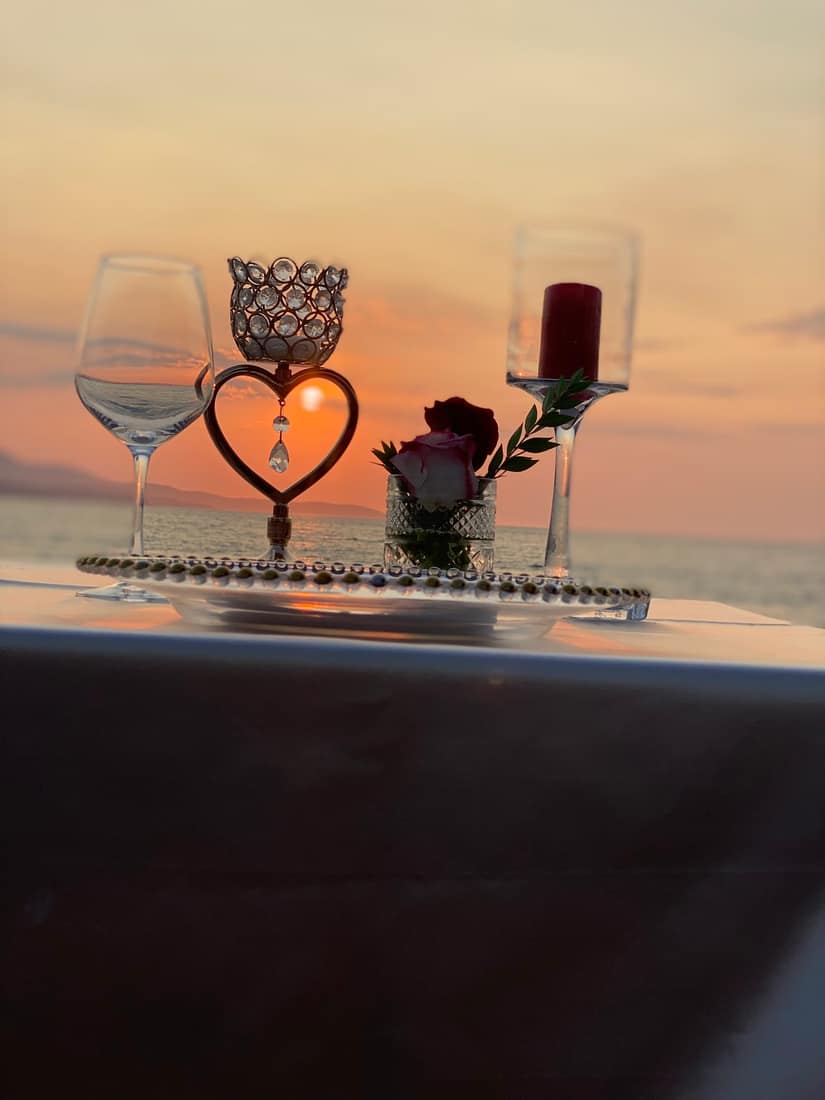 Wine glass, heart ornament and red candle on a table facing a sunset over the sea